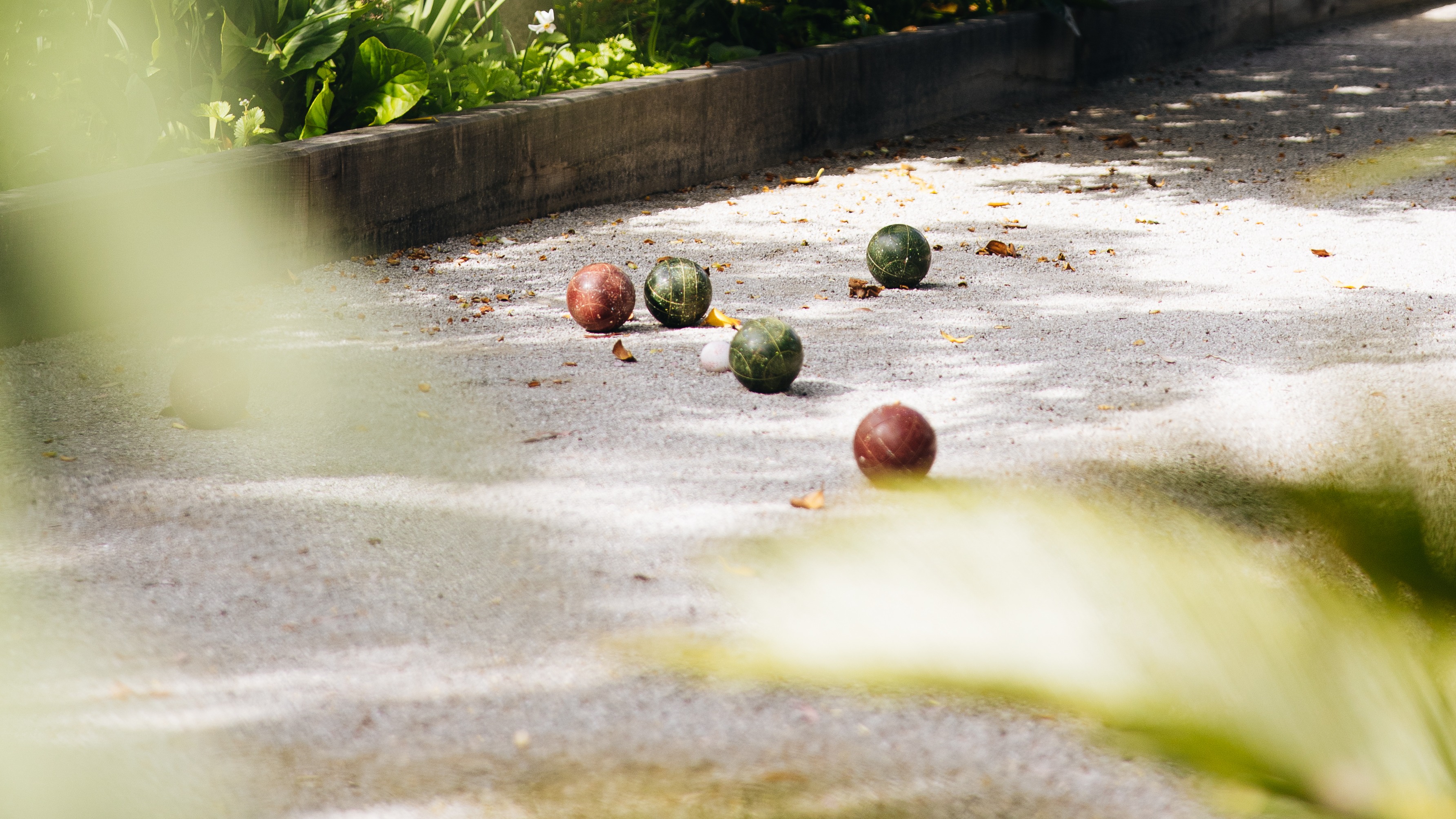Farbenfrohe Boccia-Kugeln, die auf einem Kiesplatz mit einer weißen Zielkugel verstreut sind, umgeben von grünen Gartenpflanzen.