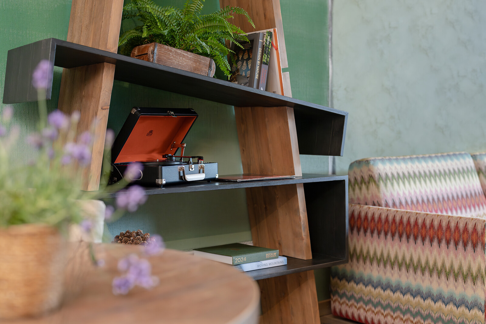 Close-up of a living area with a modern shelf, decorative plants, books, and a record player, with a patterned armchair in the background.