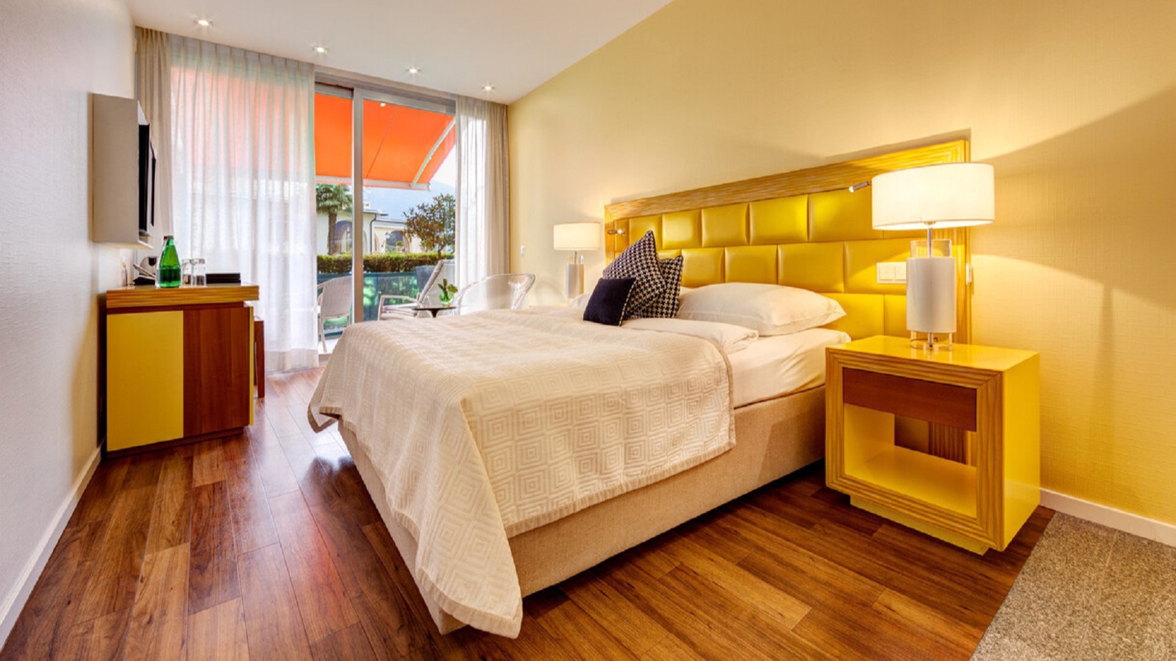 Sunny hotel room with yellow upholstered bed, matching nightstands, wooden flooring, cozy seating area, flat-screen TV, and balcony with orange awning.