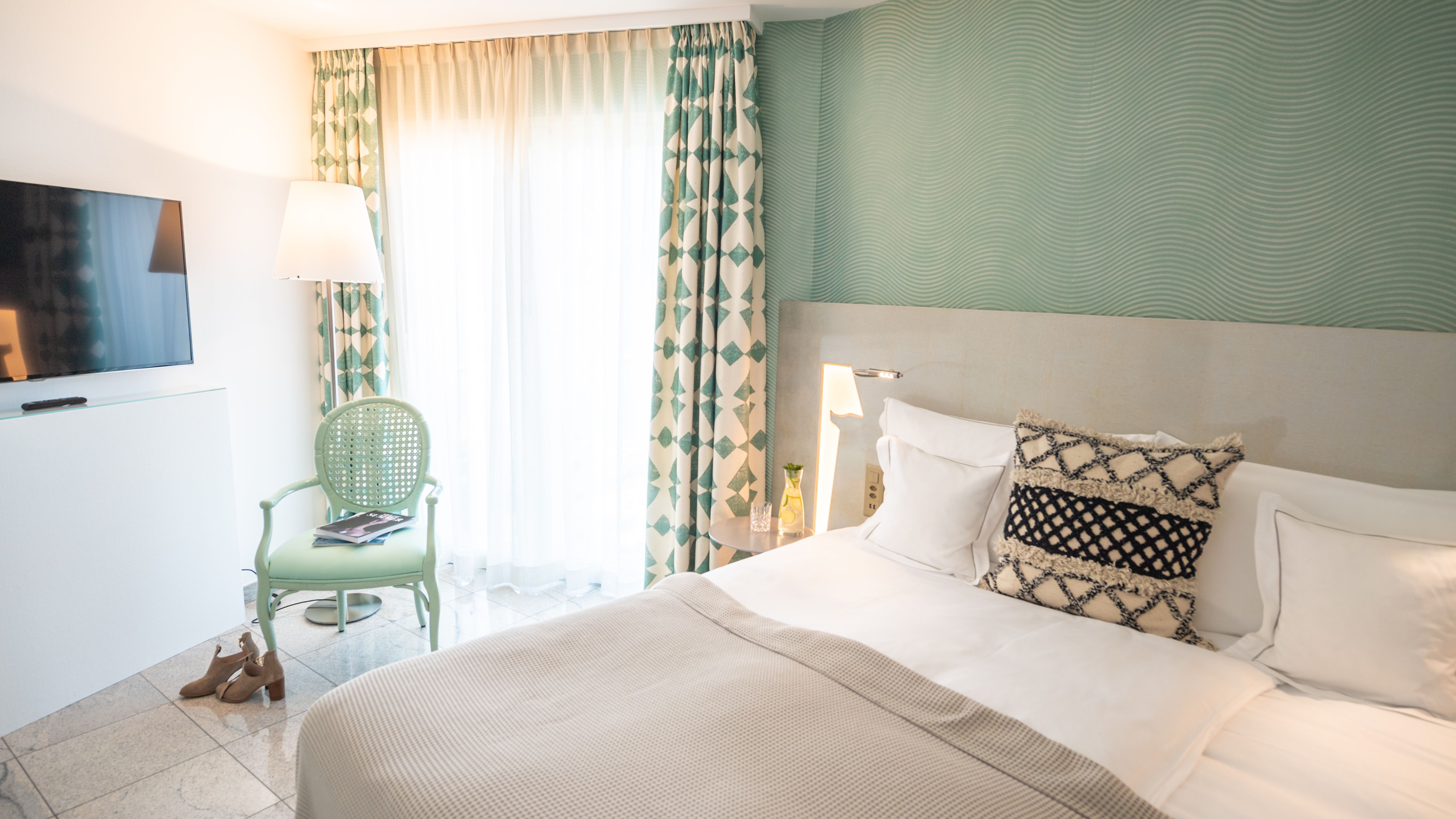 Modernes Hotelzimmer mit grüner Wellenwand, weissem Bett, mintfarbenem Sessel, gemusterten Vorhängen und einem TV.