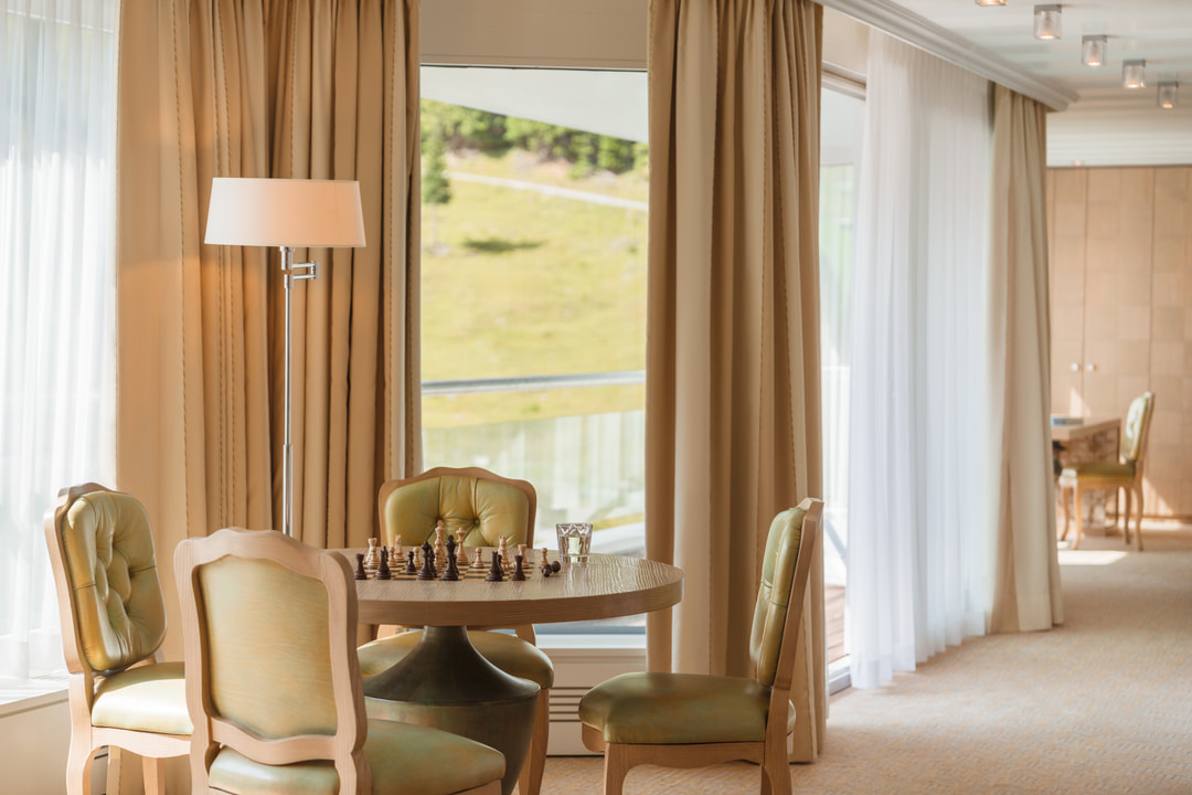 Bright hotel suite with a round table set for chess, four green upholstered chairs, floor-to-ceiling windows with beige curtains and views of the sunlit Alpine landscape.