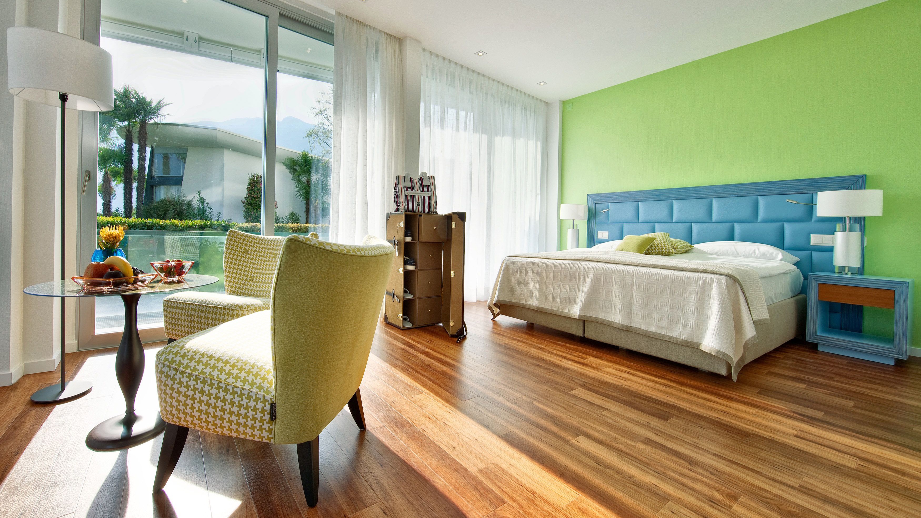 Bright hotel room with green accent wall, blue headboard, seating area with yellow armchairs, wooden floor, and garden view.