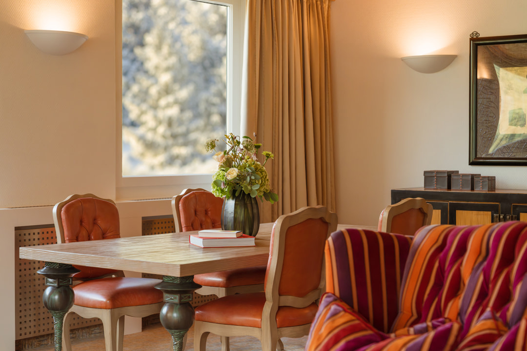Eleganter Wohnbereich eines Hotels mit Holzesstisch, vier orangefarbenen Lederstühlen und Blumenarrangement, ergänzt durch farbenfroh gestreifte Sitzmöbel, warme Wandbeleuchtung und ein grosses Fenster.