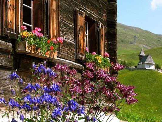 A traditional wooden house with flower-filled window boxes stands in a green mountain landscape, with a small chapel in the background.