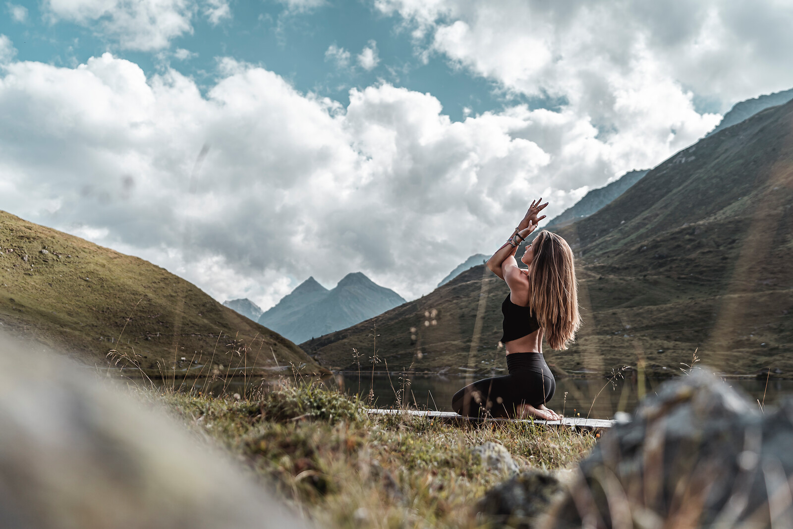 Woman practicing yoga on a mat in a mountain valley with dramatic cloudy blue sky and green hillsides in the background.