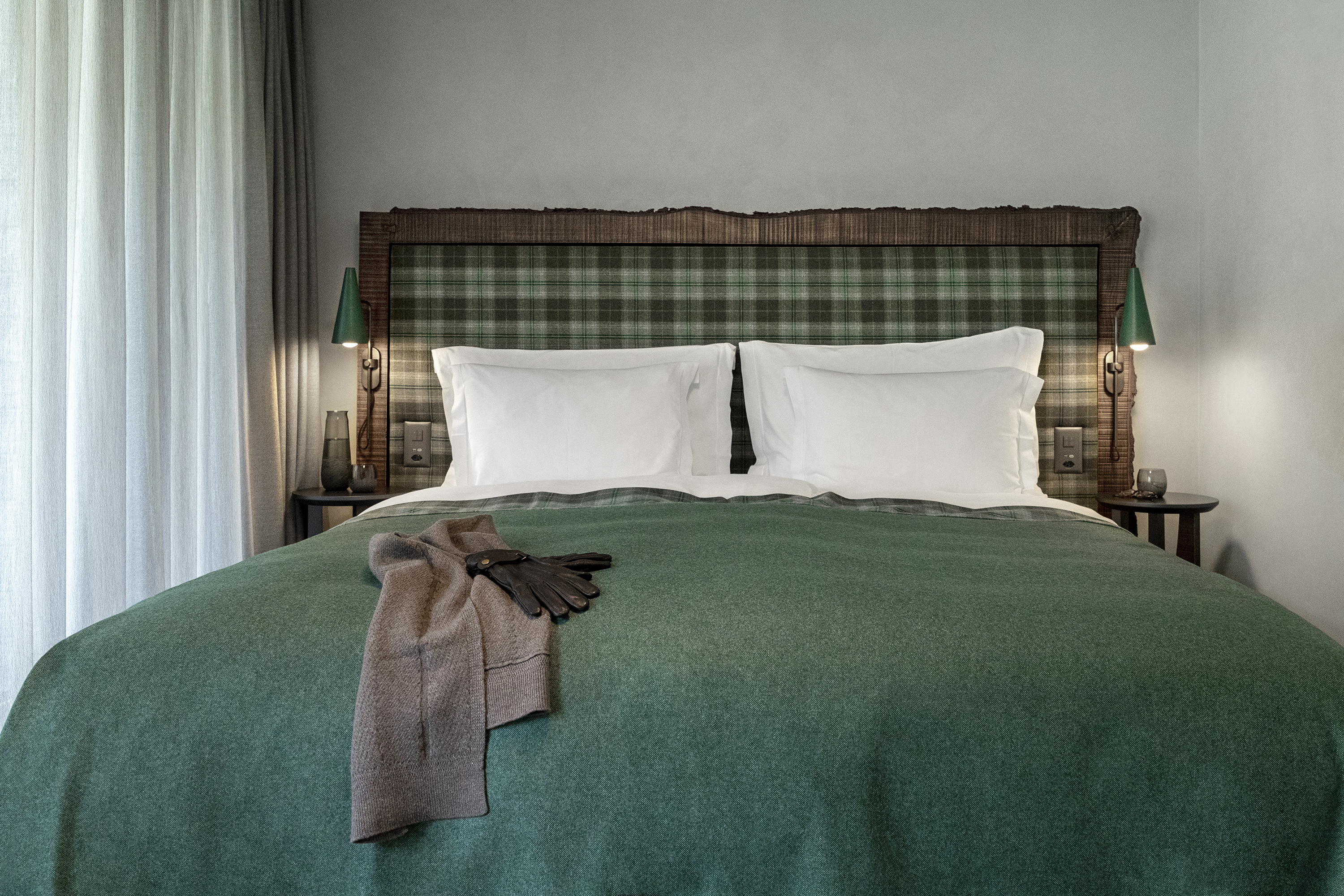 Modernes Schlafzimmer mit einem grün-karierten Kopfteil, weißen Kissen, einer grünen Bettdecke, flankierenden grünen Lampen und grauen Vorhängen.