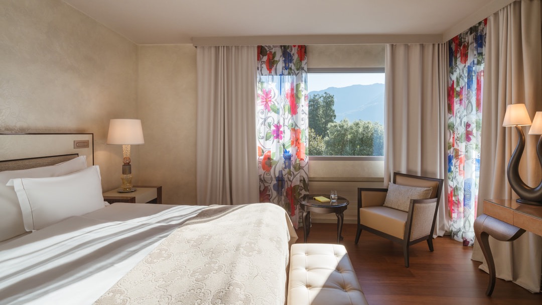 Komfortables Hotelzimmer mit floralen Vorhängen, Sitzbereich am Fenster, Schreibtisch mit Designerlampe und stilvollem Interieur in Naturtönen.