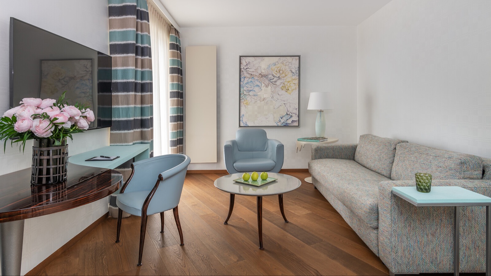 Modernes Wohnzimmer mit Flachbildfernseher, gepolsterten Sesseln in Hellblau, gestreiften Vorhängen und dekorativem Couchtisch mit grünen Äpfeln.