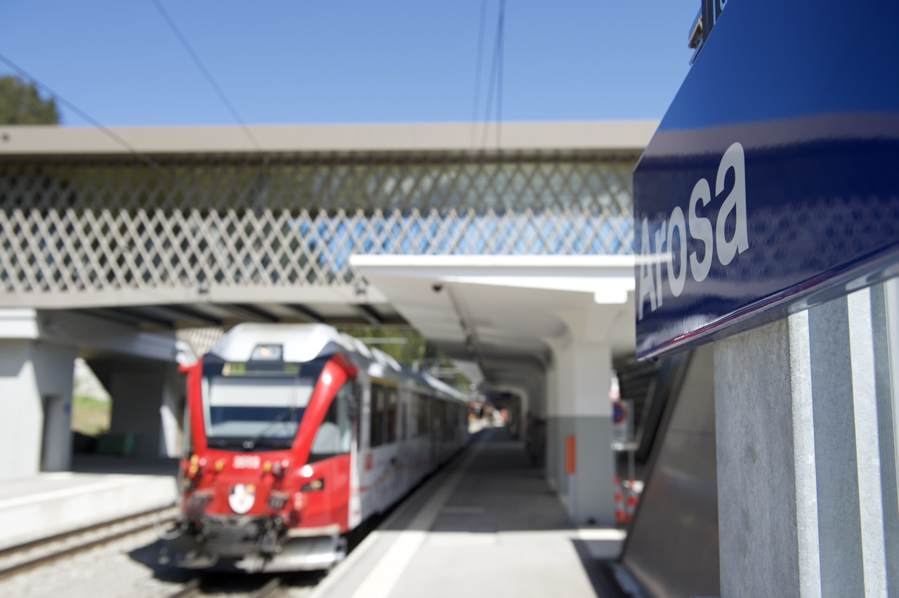 Roter Zug am Bahnhof Arosa unter blauem Himmel, Schild mit Arosa-Schriftzug im Vordergrund.