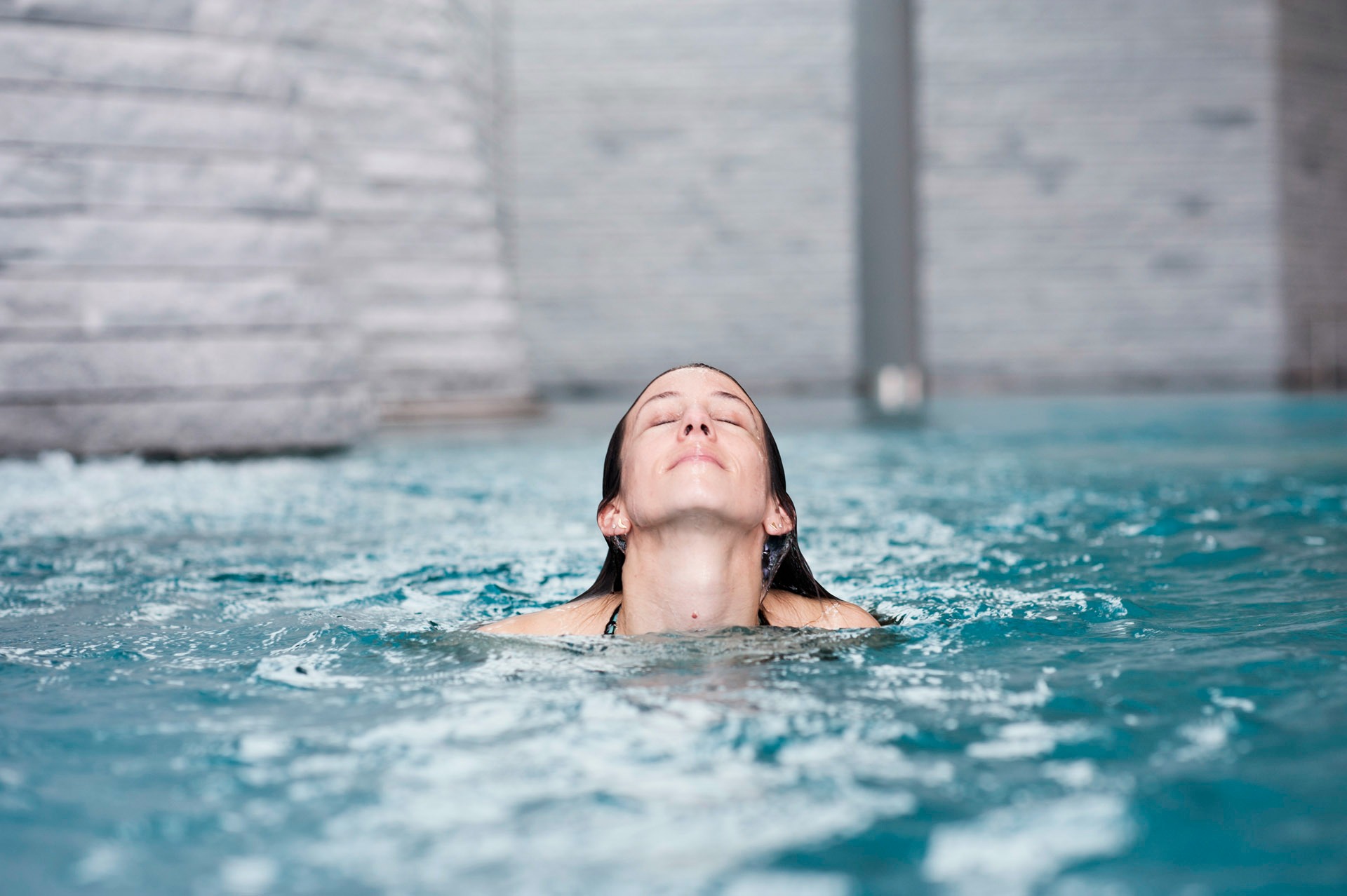 Eine Frau entspannt mit geschlossenen Augen im türkisfarbenen Poolwasser, den Kopf in einem friedlichen Moment der Ruhe nach hinten gelehnt.