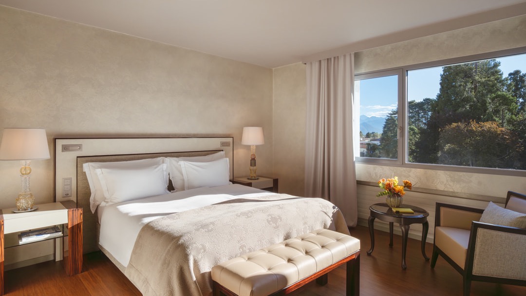 Helles Hotelzimmer mit Kingsize-Bett, eleganter Sitzbank, Naturholzfußboden, zwei Nachttischen und großem Fenster mit Blick auf Bäume und Berge.