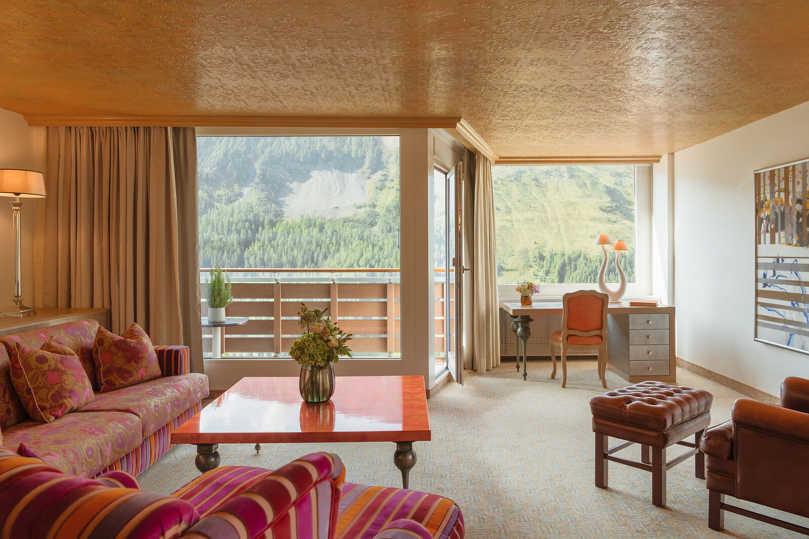 Elegant hotel living room with colourful patterned sofas, a red coffee table, leather seating and a writing desk, framed by large windows and a balcony with sweeping Alpine mountain views