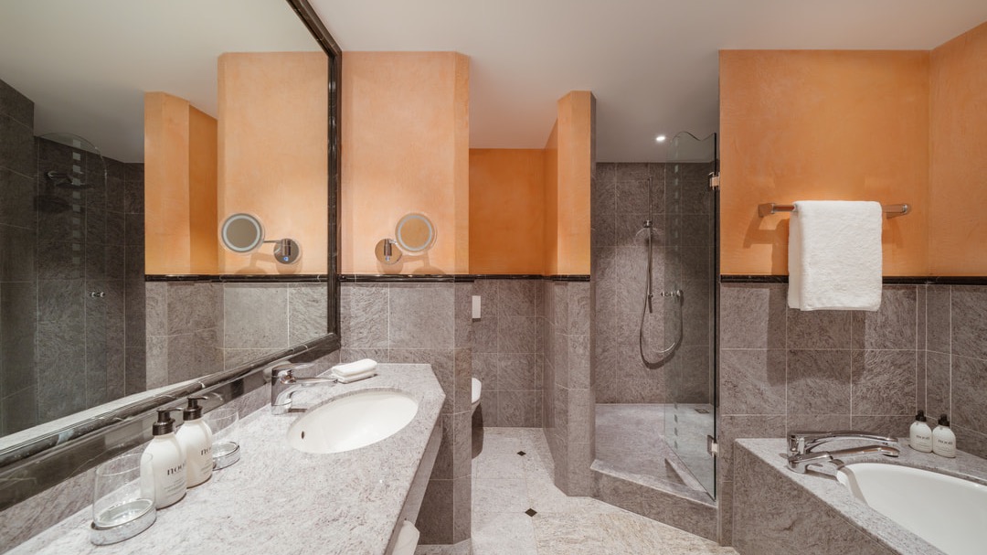 Modernes Badezimmer mit Waschbecken, Badewanne, geräumiger Dusche, Wandspiegeln und orange-grauer Farbgestaltung.