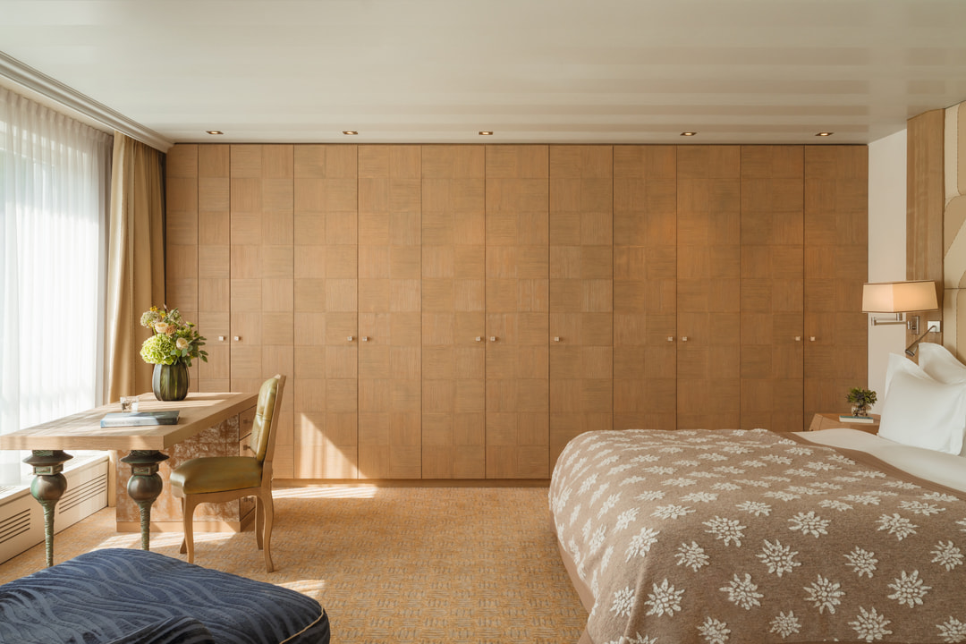 Bright, elegant hotel bedroom with floor-to-ceiling windows, a large bed with a patterned beige bedspread, a wooden writing desk with an upholstered chair and extensive built-in wooden wardrobes.