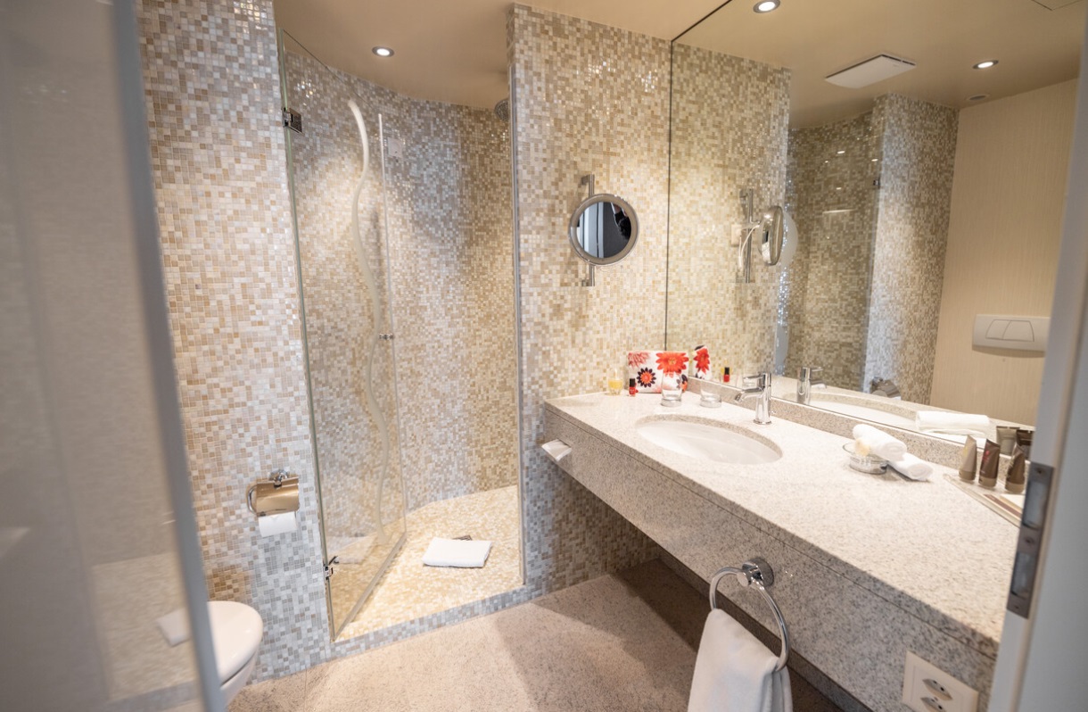 Modernes Badezimmer mit beigefarbenen Mosaikfliesen, gläserner Dusche, Waschtisch mit Spiegel und eingebauter Beleuchtung.