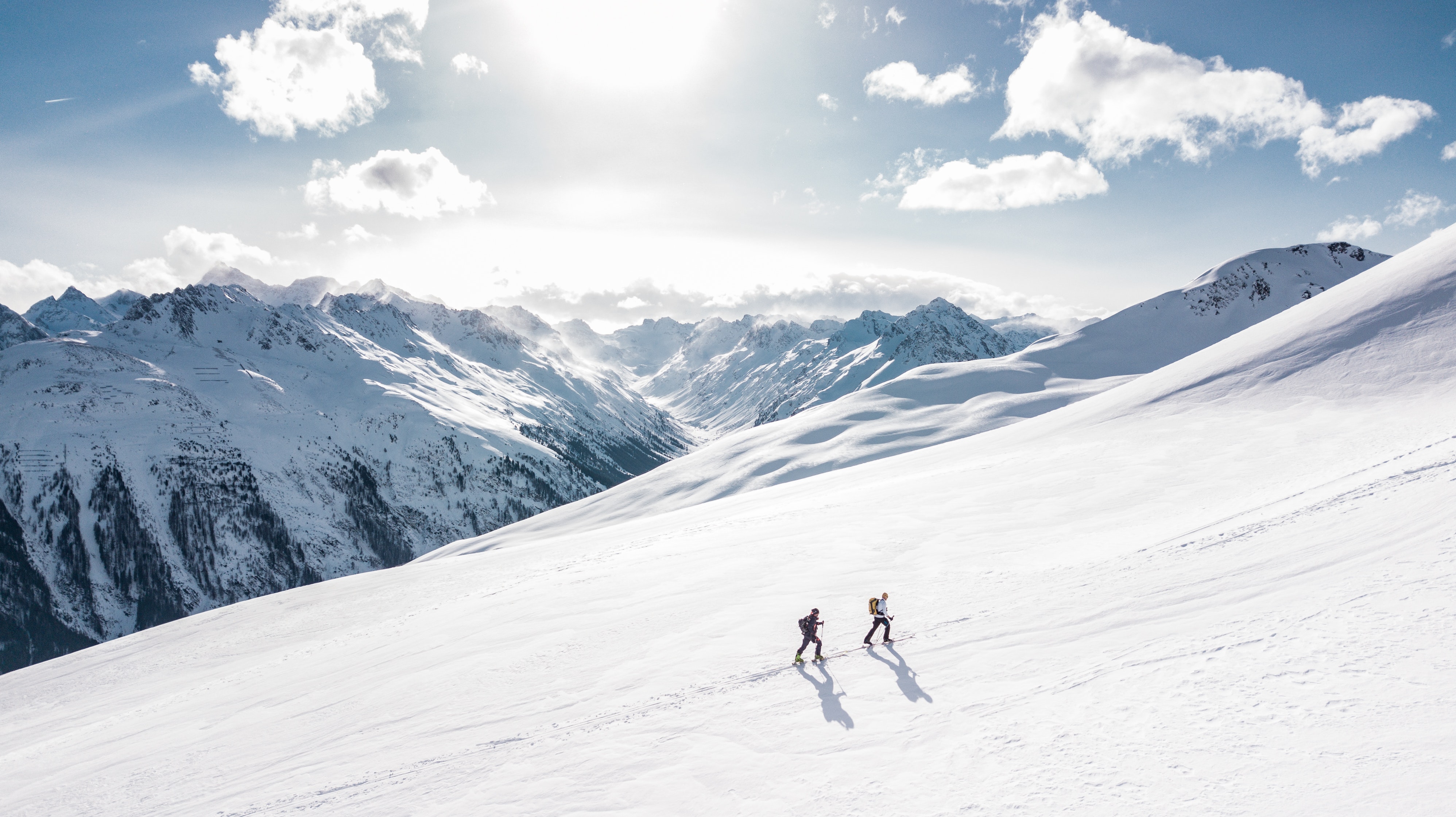Zwei Skifahrer beim Aufstieg an einem unberührten, schneebedeckten Berghang vor einer Kulisse aus dramatischen Gipfeln und weißen Wolken unter einem strahlend blauen Himmel.