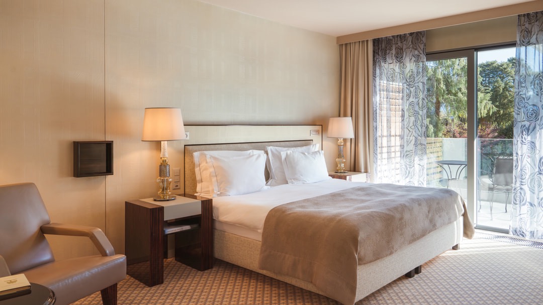 Bright hotel room with double bed, two bedside tables with lamps, beige bedspread, floor-to-ceiling floral curtains, and balcony overlooking the garden.