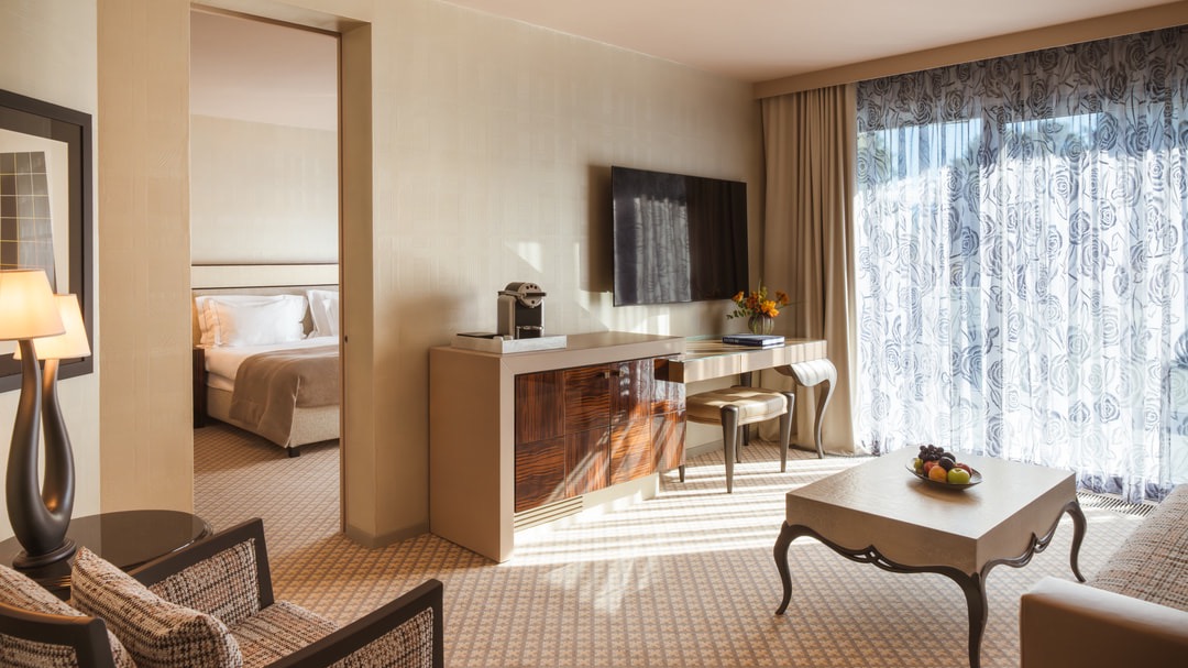 Modern living area featuring a flat-screen TV, desk with chair, Nespresso machine, coffee table with fruit bowl, and access to the bedroom.