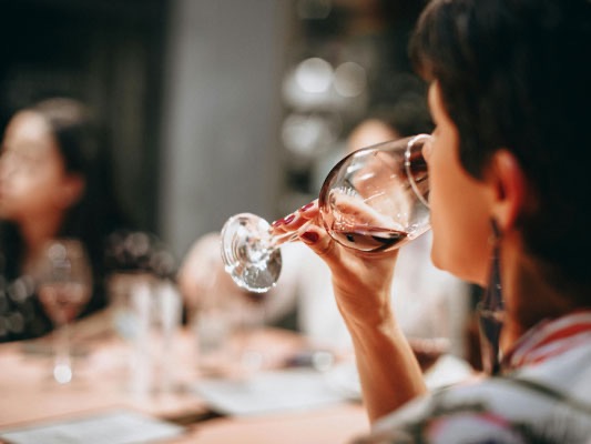 A woman tastes red wine from a glass during a wine tasting, with other guests seated at the table in the background.