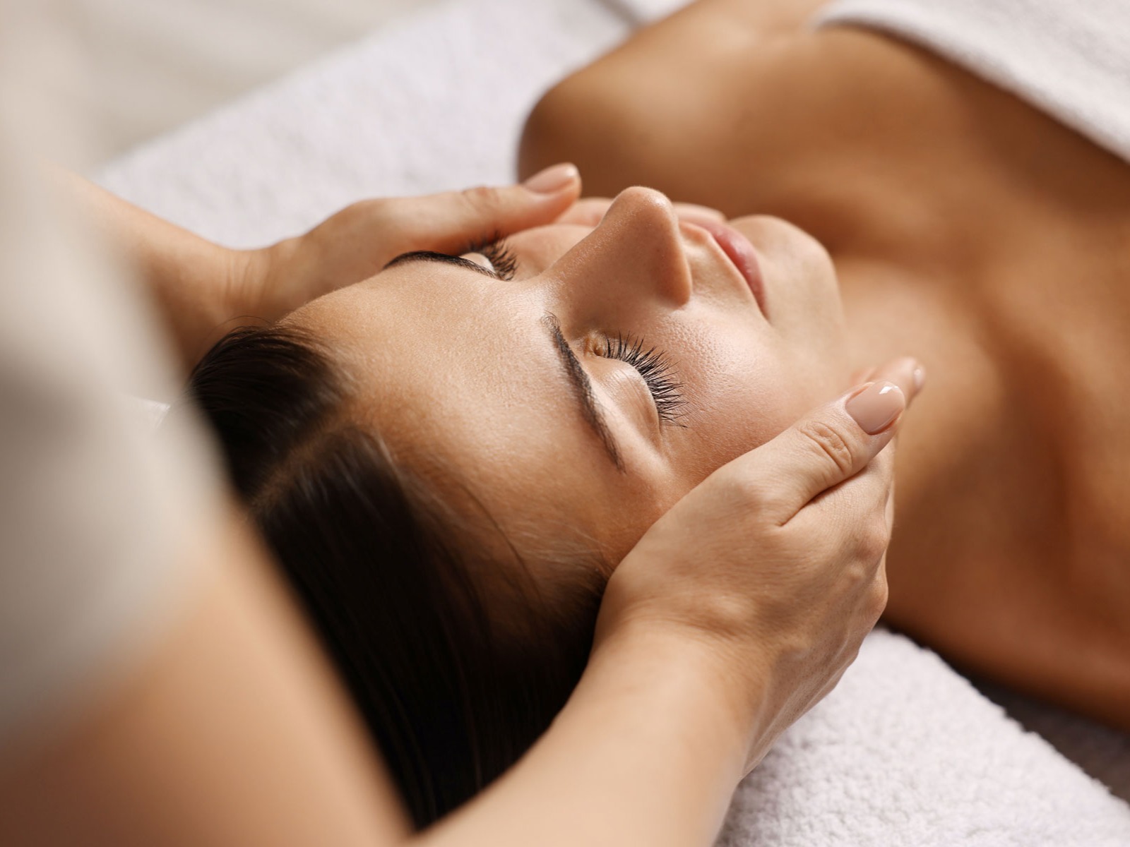 A woman lies with her eyes closed on a spa bed while her face is gently held with both hands during a relaxing facial massage.