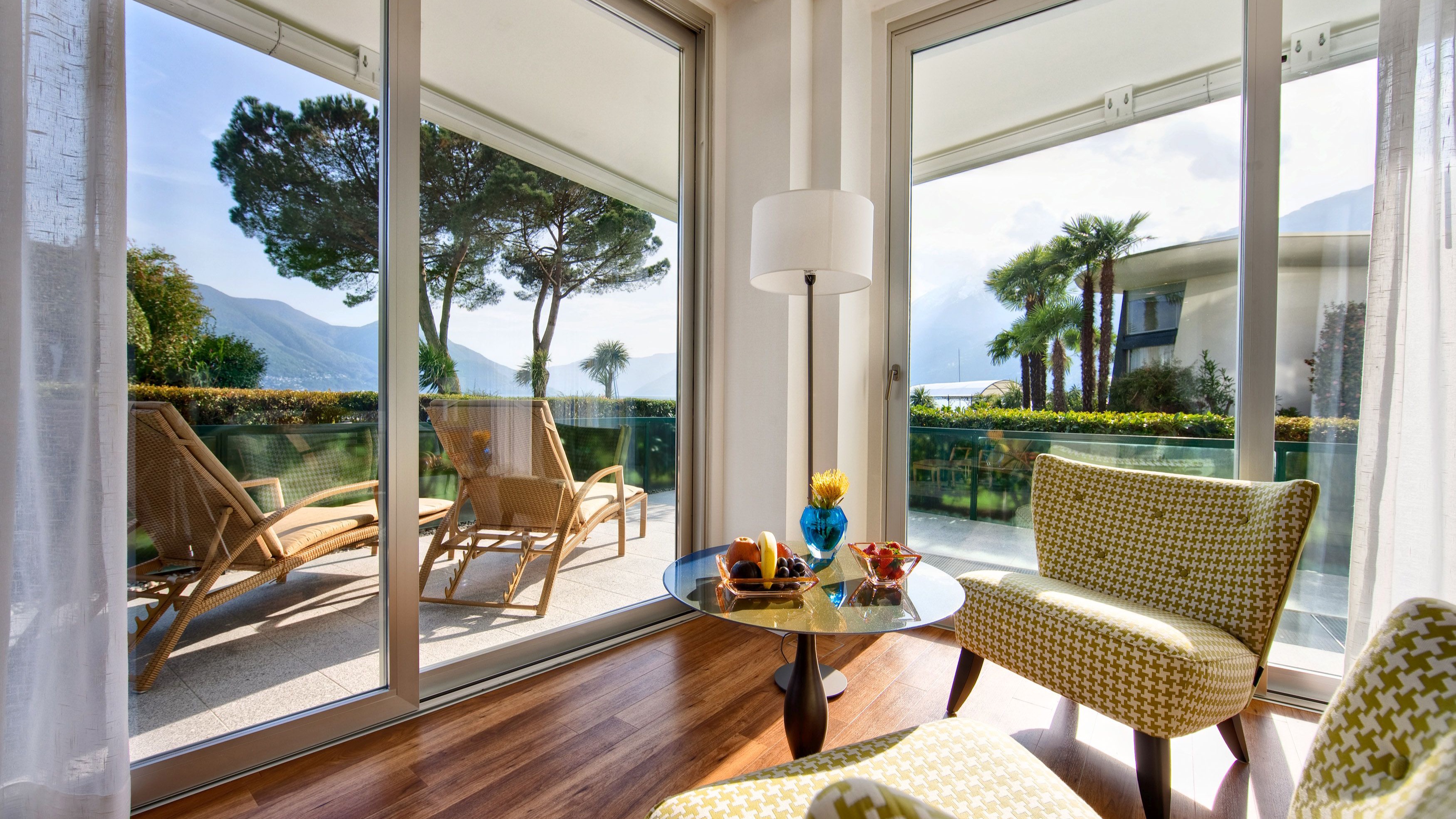 Sunny lounge area with glass table, yellow chairs, snacks, and panoramic view of terrace with palm trees and mountains.