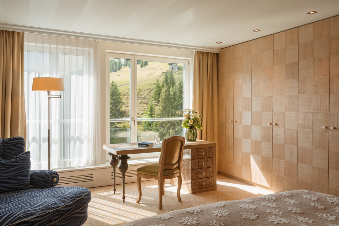 Helles Hotelzimmer mit Holzschreibtisch und gepolstertem Stuhl vor grossen Fenstern mit Blick auf einen grünen Alpenhang, dazu Einbauschränke aus Holz, ein dunkelblauer Sessel und ein Bett mit gemusterter beiger Tagesdecke.