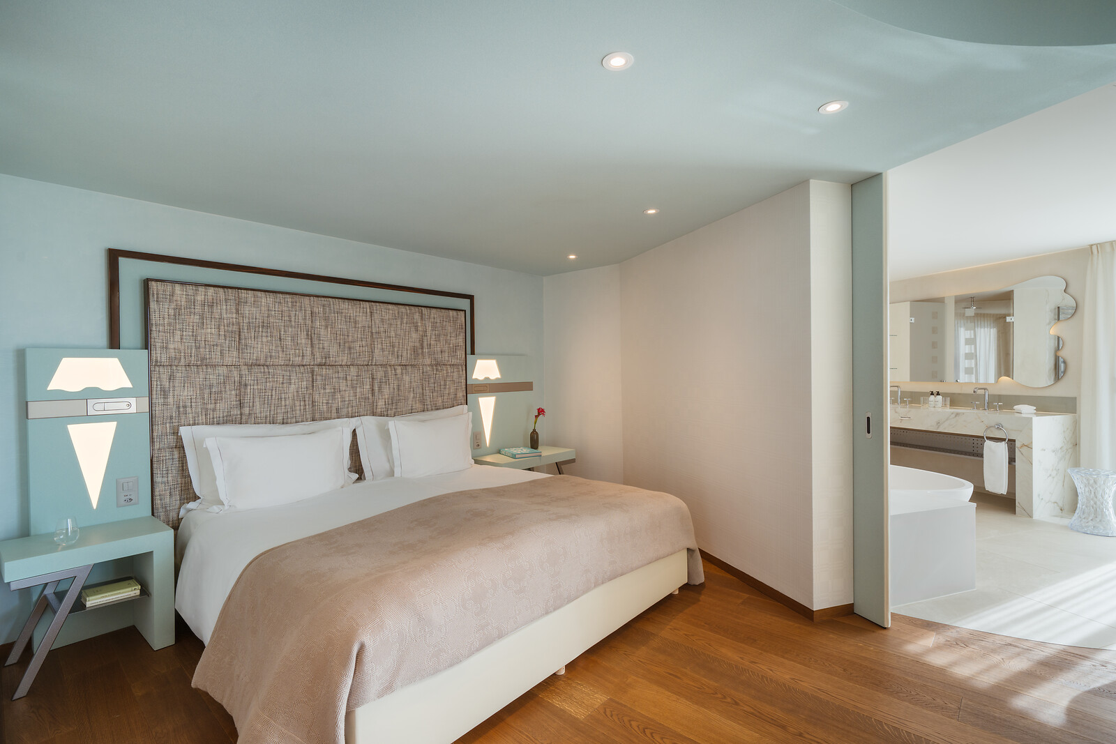 Stilvolles Hotelzimmer mit modernem Doppelbett, hellblauem Interieur, weichem Holzboden und offenem Übergang zum luxuriösen Badezimmer mit freistehender Badewanne und Doppelwaschtisch.