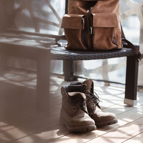 Braune Lederstiefel stehen vor einer Bank mit braunem Rucksack in sonnendurchflutetem Raum.