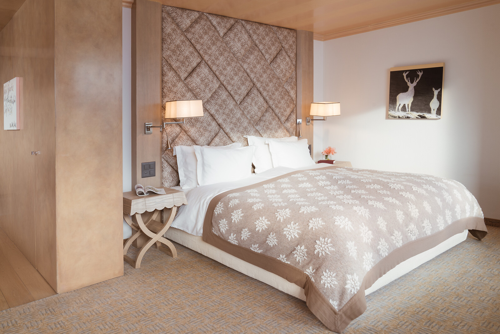 Elegant hotel bedroom with a large double bed, a textured woven headboard, crisp white pillows and a beige floral bedspread, complemented by warm wall lamps, wooden furnishings and deer artwork.