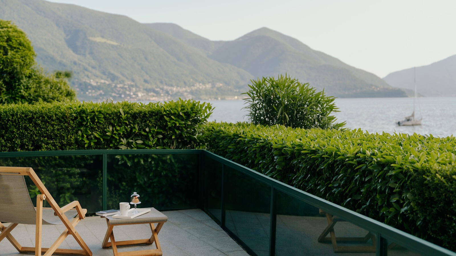 Terrasse mit Liegestuhl und Beistelltisch vor grüner Hecke, Blick auf See und Berge im Hintergrund