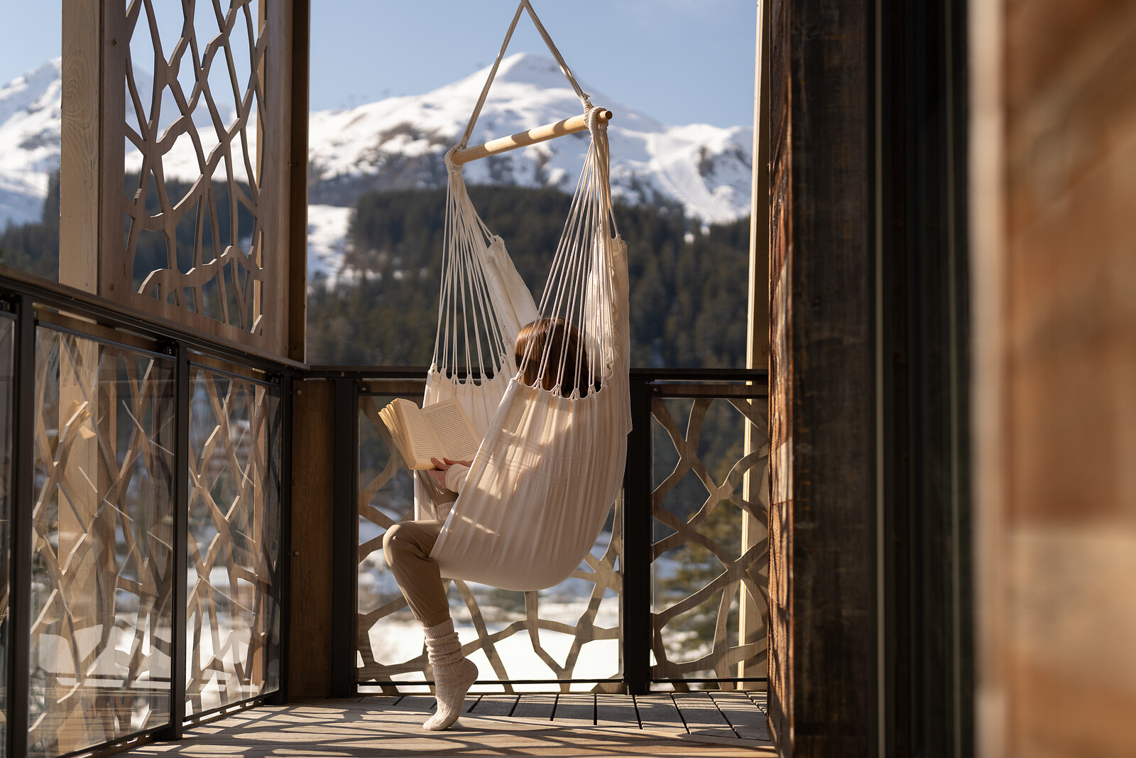 Eine Person entspannt in einem Hängesessel auf dem sonnigen Balkon des Valsana Hotels und liest ein Buch, mit Blick auf die verschneite Berglandschaft von Arosa.