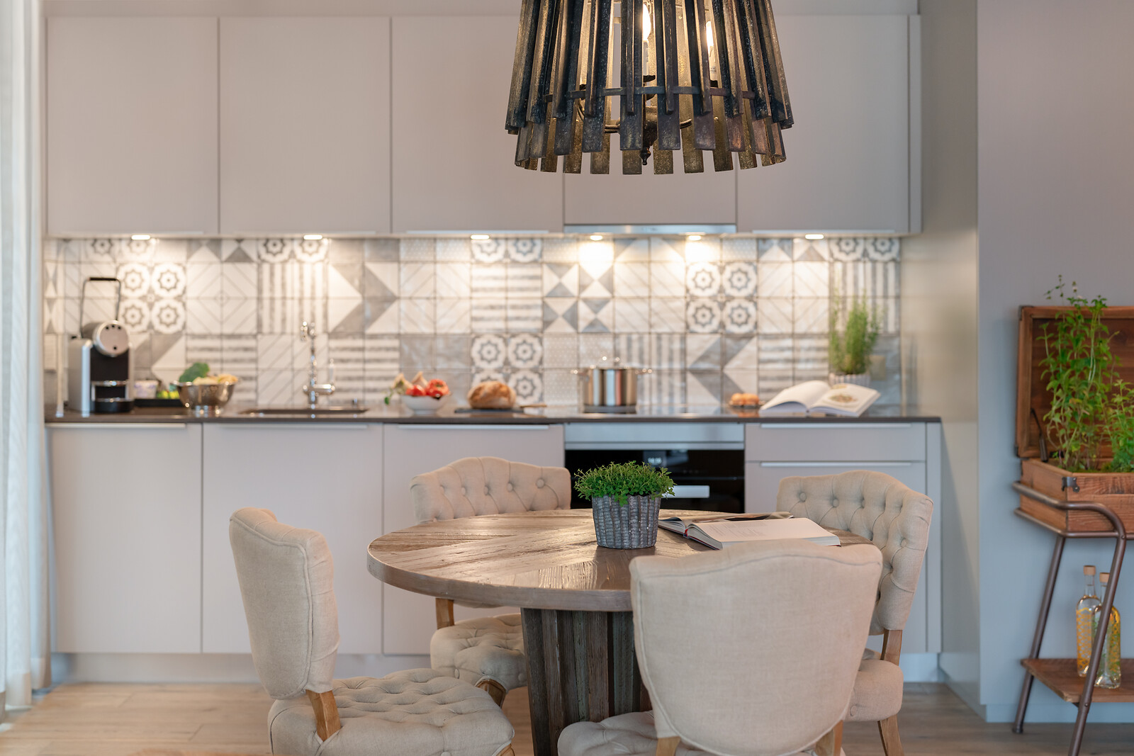 Modern kitchen and dining area with a wooden table, upholstered chairs, stylish pendant light, and open kitchen with decorative tiled backsplash.