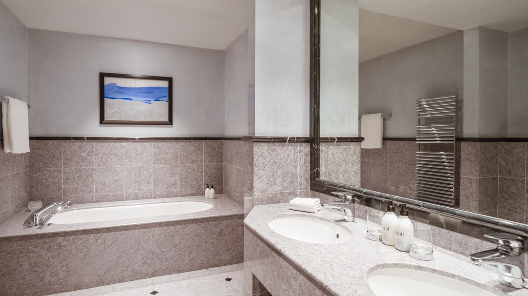 Modernes Bad mit eingelassener Badewanne, großem Wandspiegel, Doppelwaschbecken, hellgrauen Natursteinfliesen und Kunstwerk mit blauen Farbakzenten.