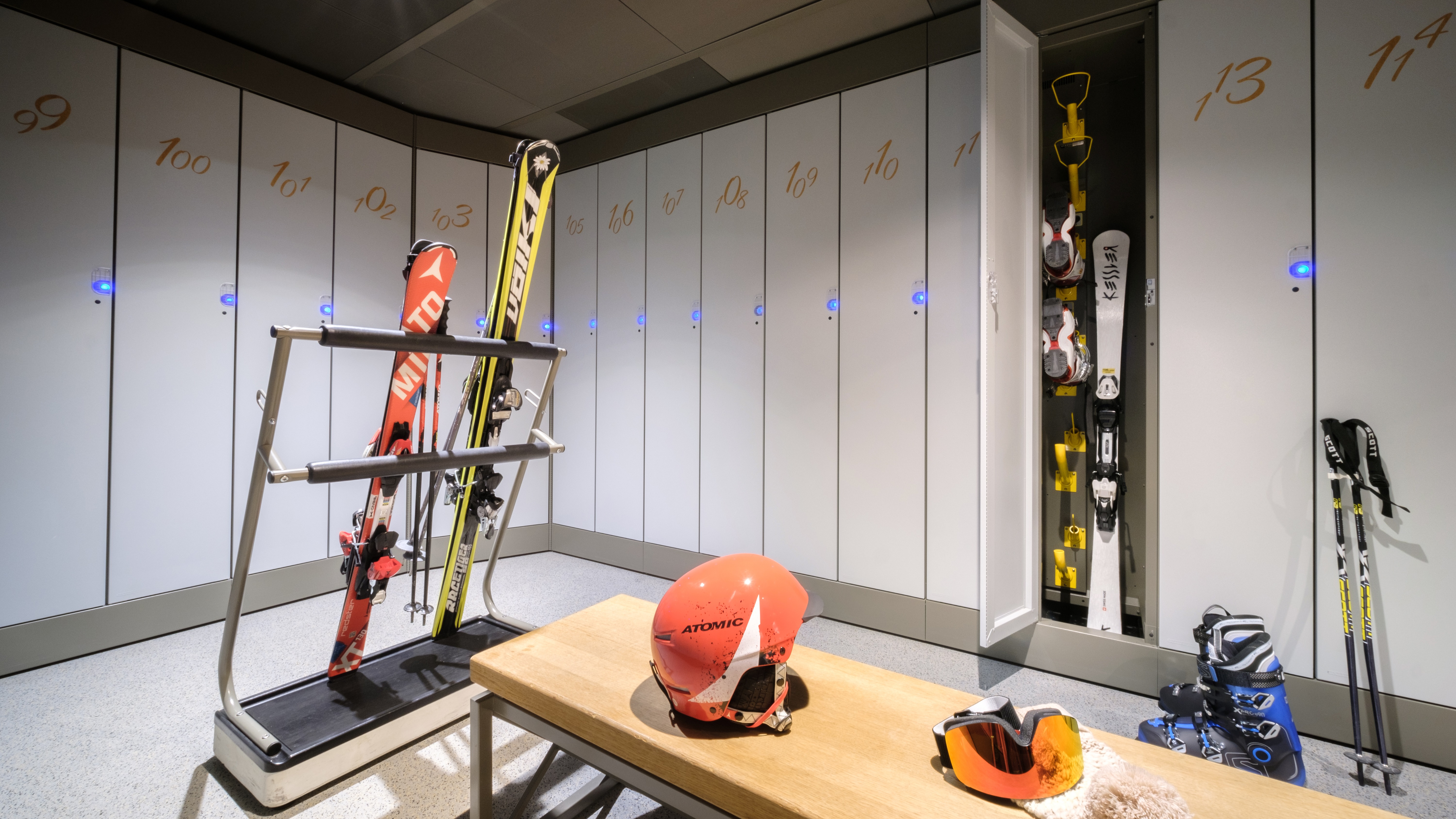 Moderner Skikeller mit nummerierten weißen Schließfächern, blauen LED-Lichtern, Skiern im Ständer, einem orangefarbenen Helm auf einer Bank und Skischuhen.