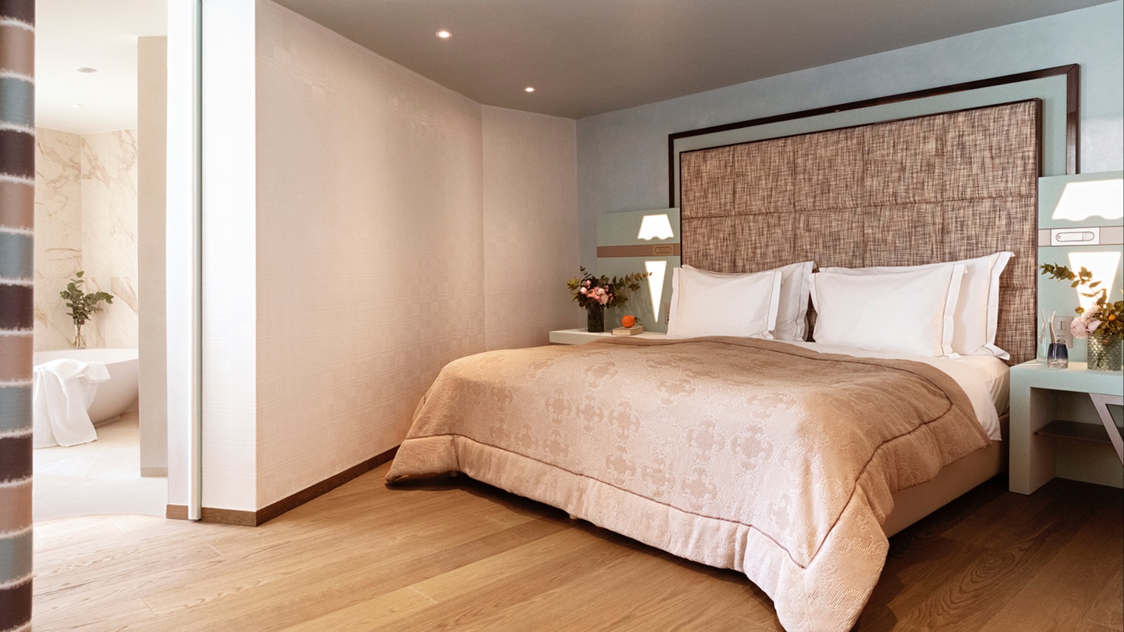 Modern hotel room with stylish beige bed, warm wooden flooring, floral arrangements, and open access to the luxury bathroom.