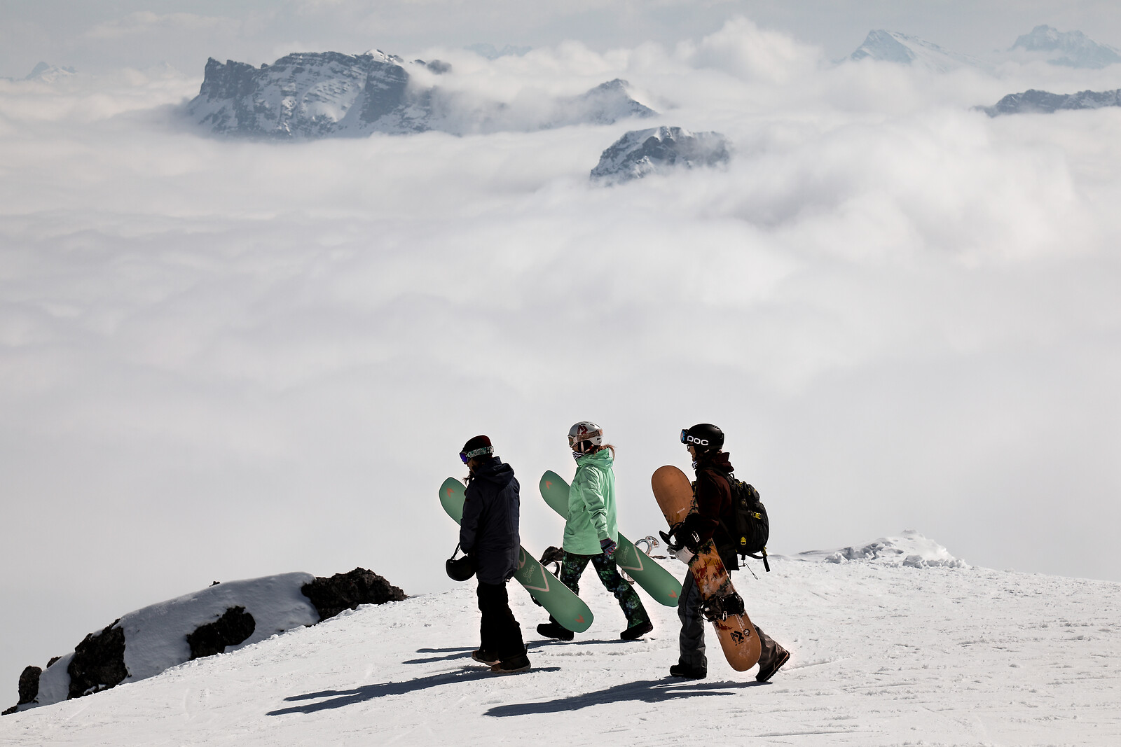 Three snowboarders walking on snowy mountain slope with clouds and rocky peaks in background