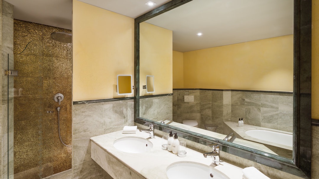 Luxuriöses Badezimmer mit großer goldener Regendusche, Doppelwaschtisch aus Stein, Wandspiegel, beigen Fliesen und Pflegeprodukten.