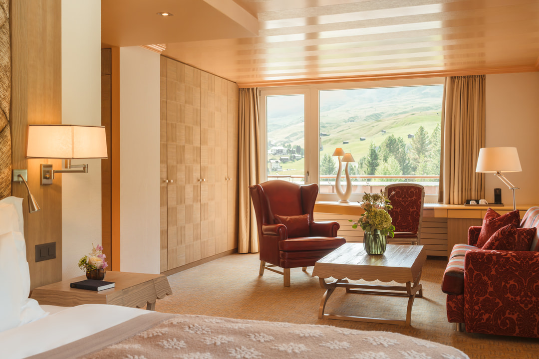 Grosszügiges Hotelzimmer mit Wohnbereich, grossem Bett, rotem Ledersessel, gemustertem Sofa und Holzcouchtisch, umrahmt von grossen Fenstern mit Blick auf die grüne Alpenlandschaft.