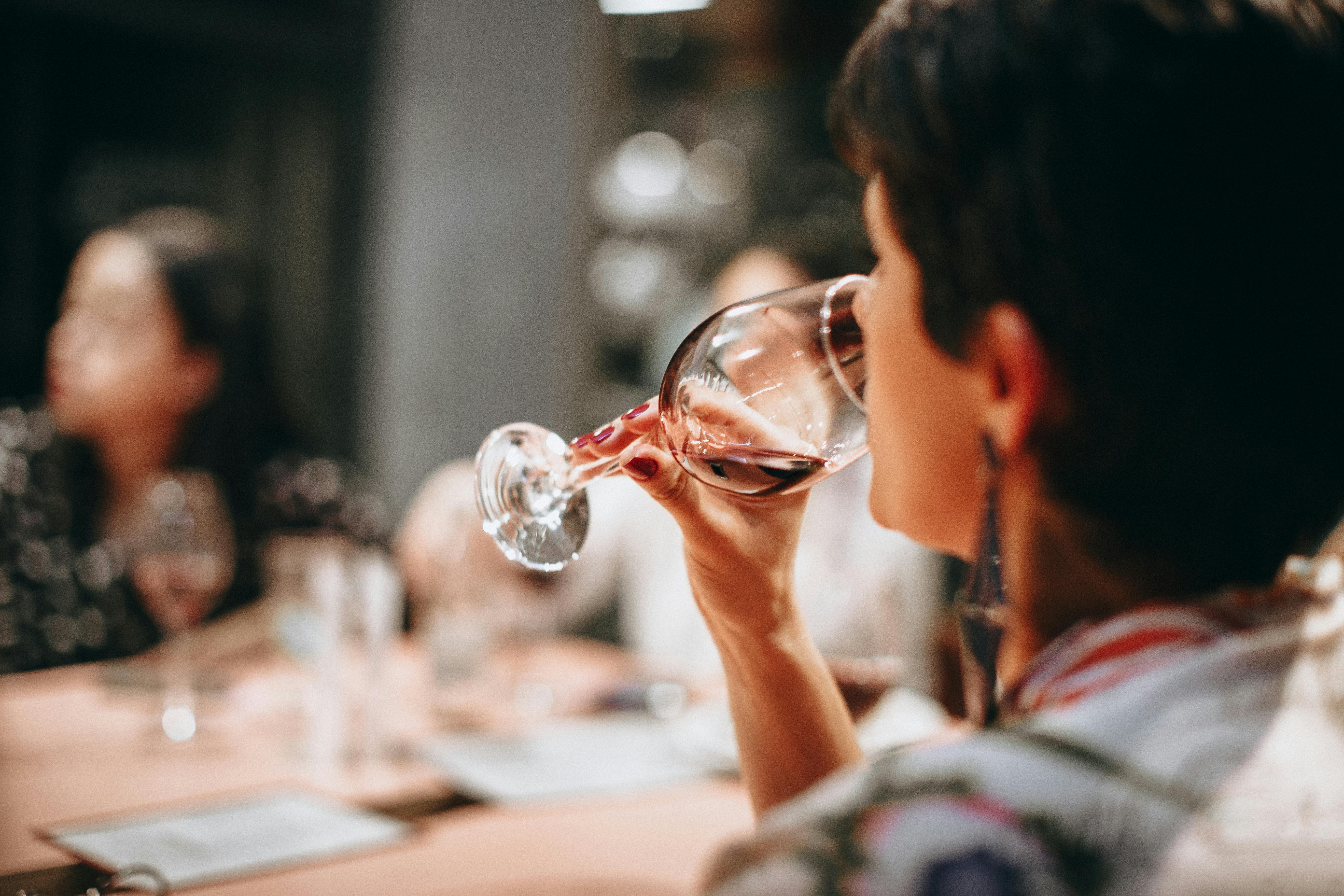 Person sipping red wine from a glass at a dining table with blurred guests in the background.