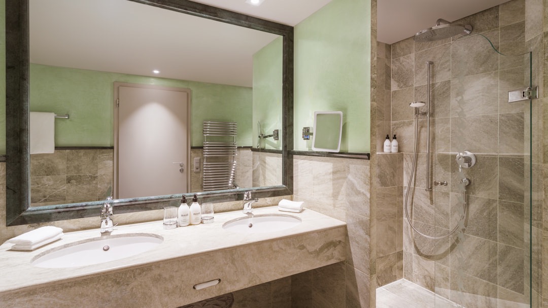 Luxuriöses Badezimmer mit großer begehbarer Regendusche, grüner Wandfarbe, Marmorfliesen, großem Spiegel und Doppelwaschtisch mit Pflegeprodukten.