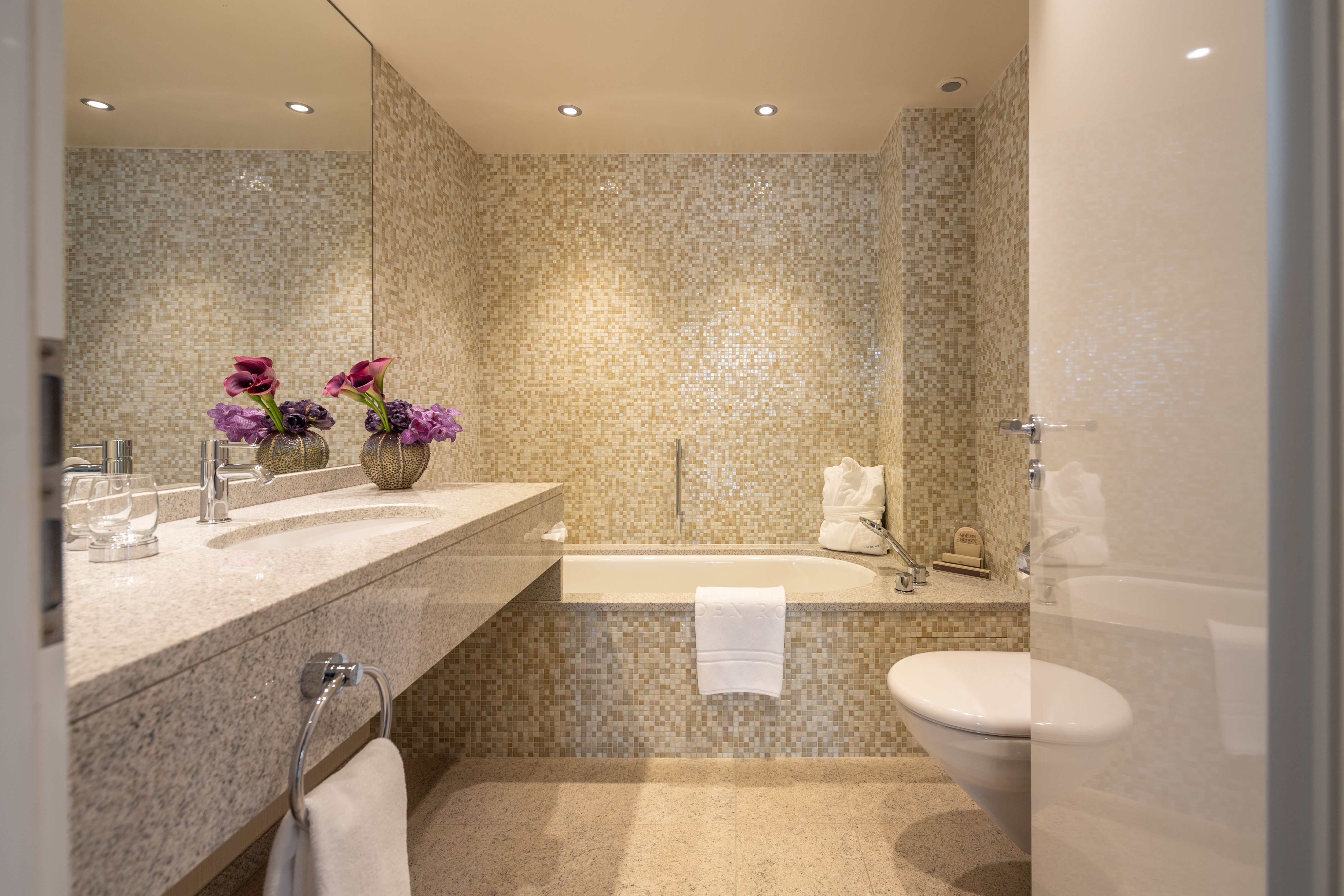 Modernes Badezimmer mit beigen Mosaikfliesen, eingebauter Badewanne, Waschbecken und lila Blumendekoration.