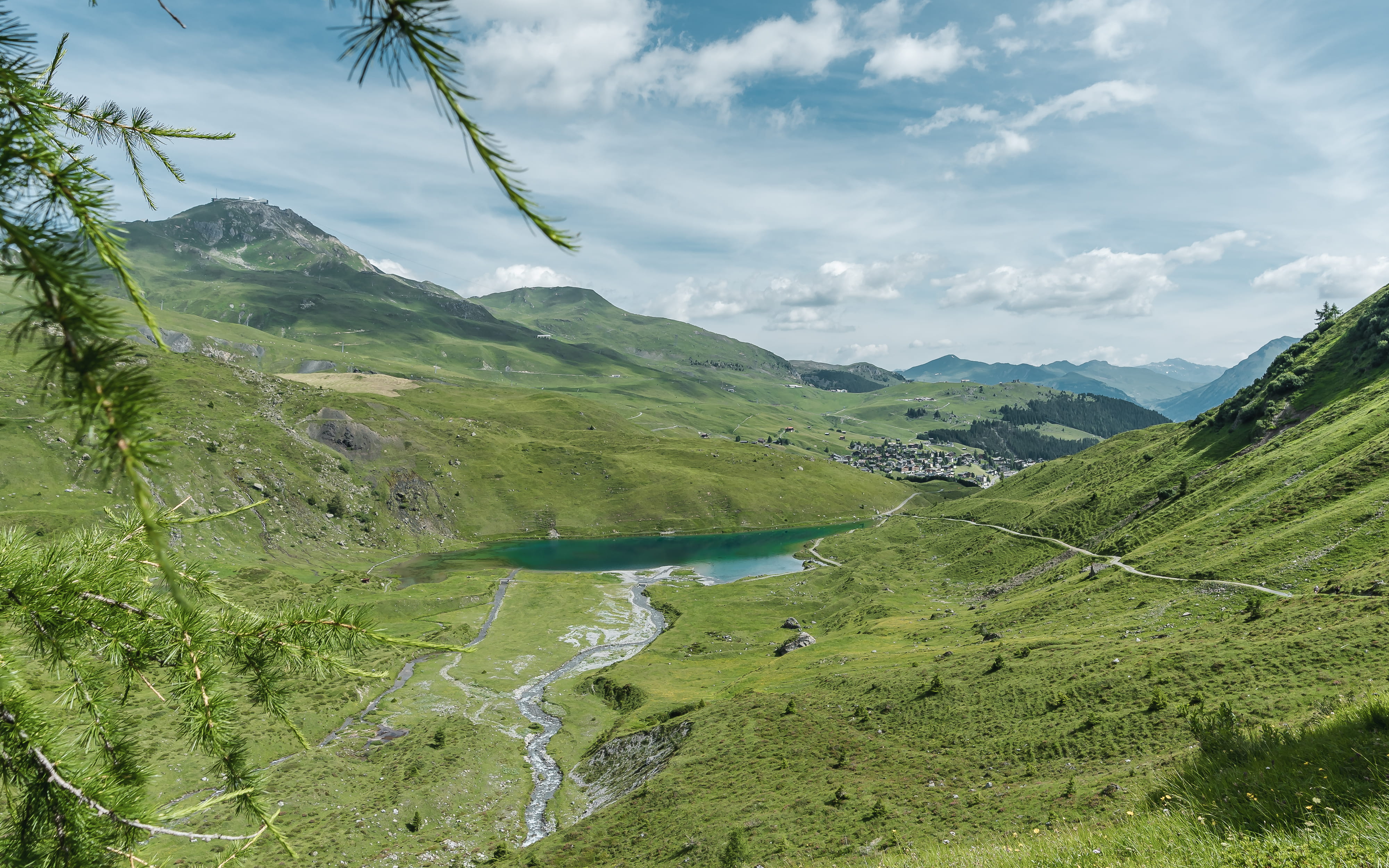 Alpiner Bergsee umgeben von grünen Hügeln, Bach fliesst ins Tal, Bergdorf im Hintergrund, blauer Himmel mit Wolken.
