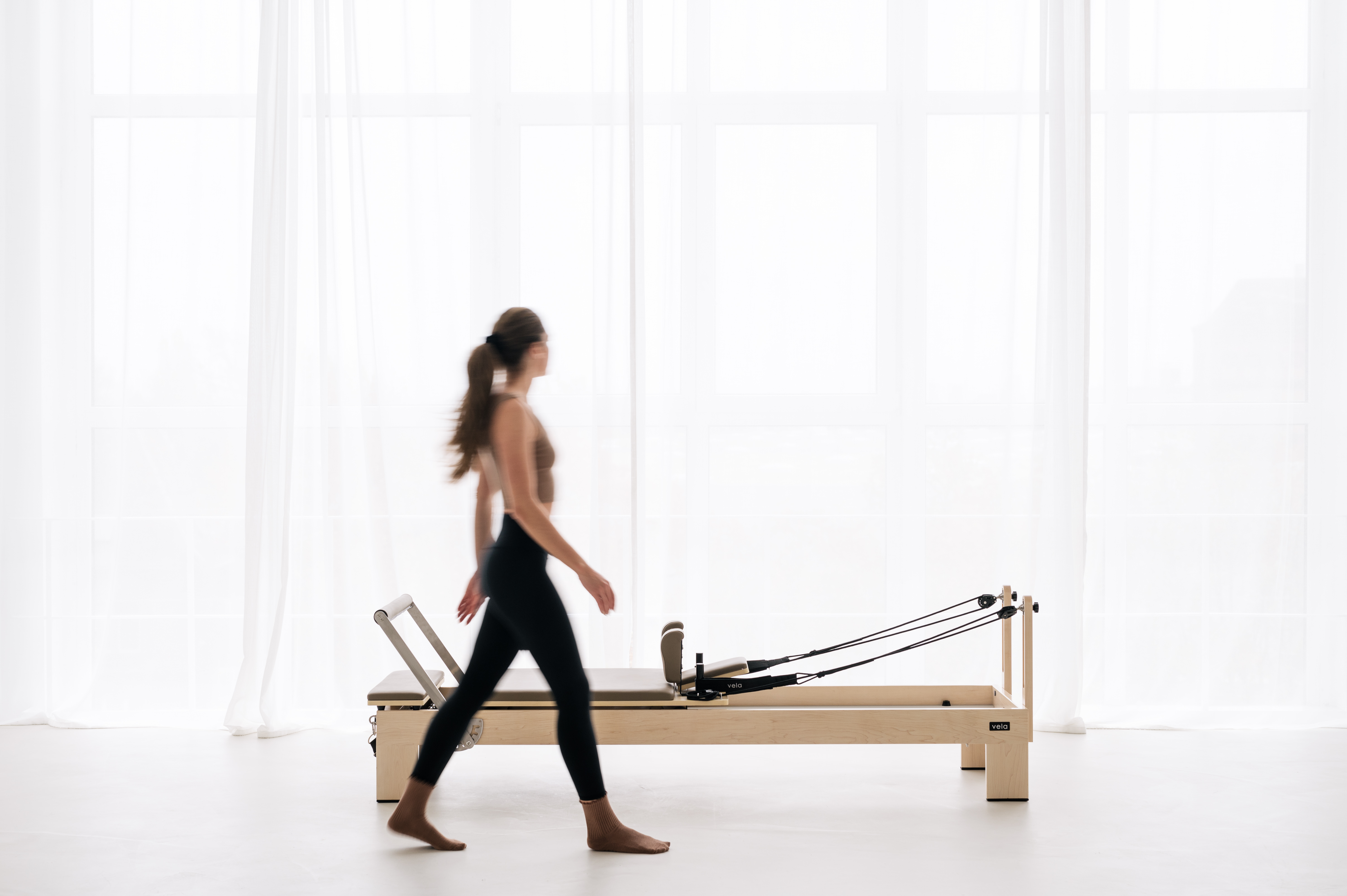 A woman walks past a Pilates reformer in a bright, light-filled studio.
