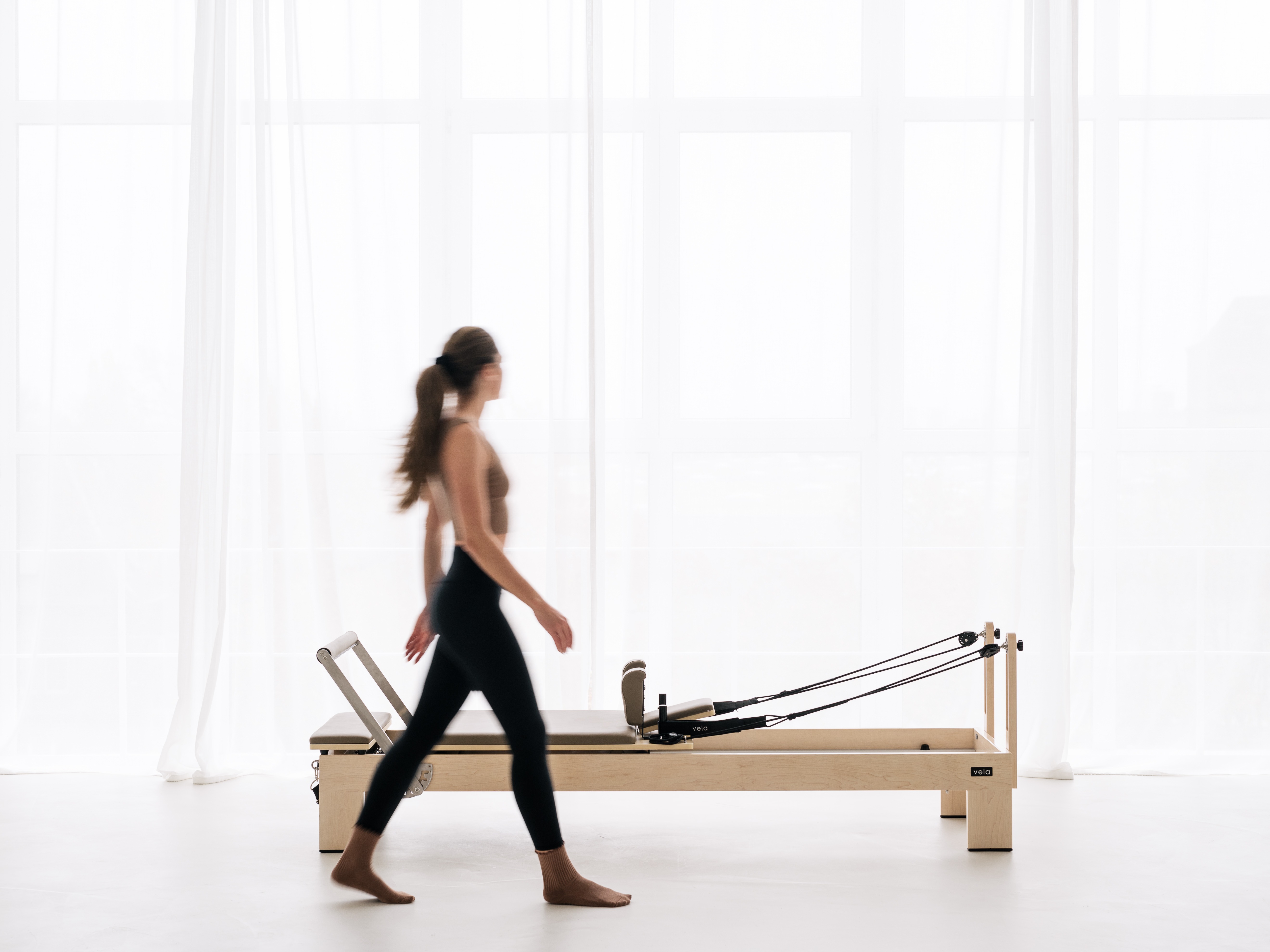 Eine Frau geht in einem hellen, lichtdurchfluteten Pilates-Studio an einem Reformer vorbei.