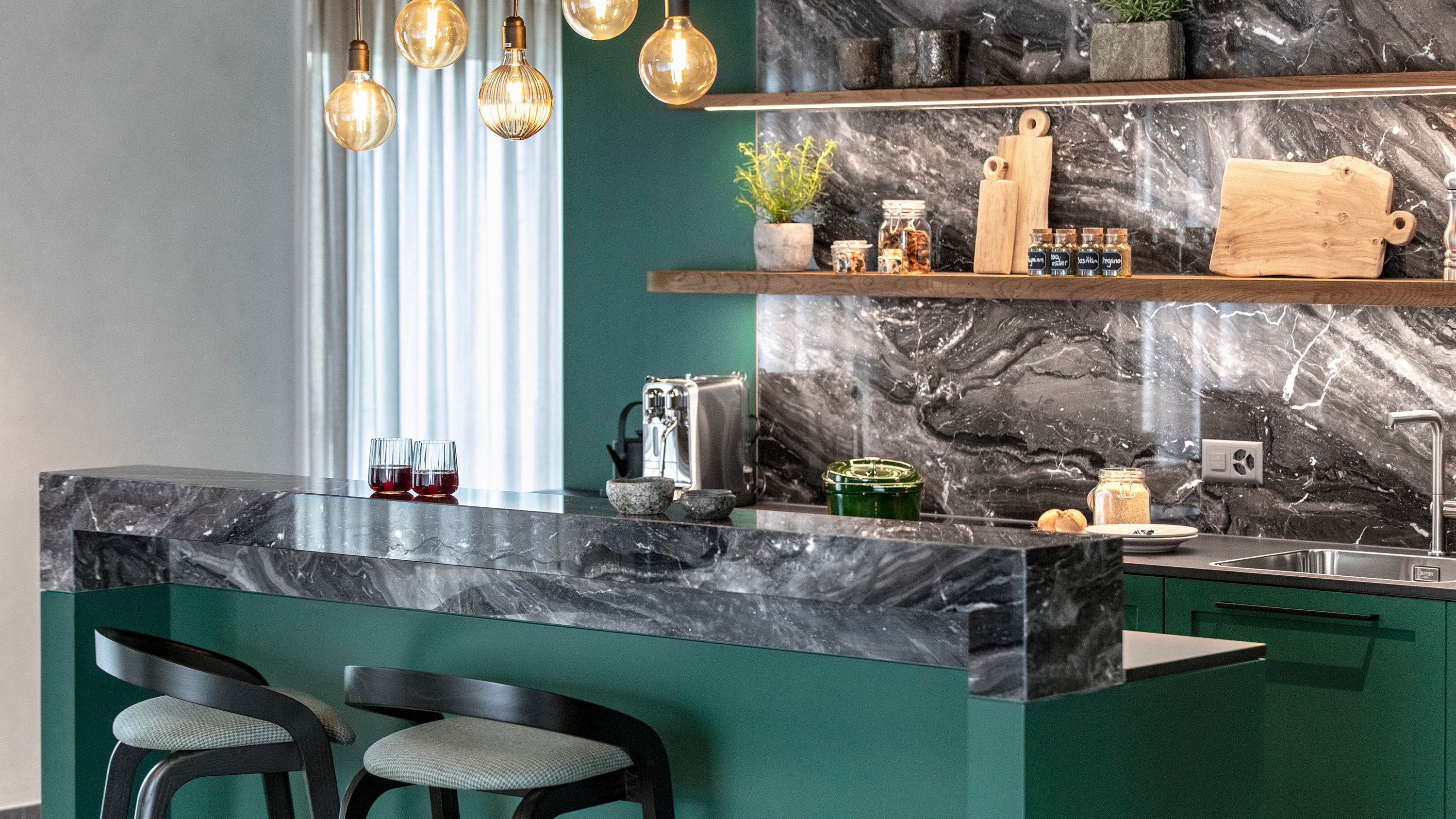 Moderne Küche mit grünen Schränken, Arbeitsplatten und Rückwand aus schwarzem Marmor, Holzregalen und bernsteinfarbenen Pendelleuchten.