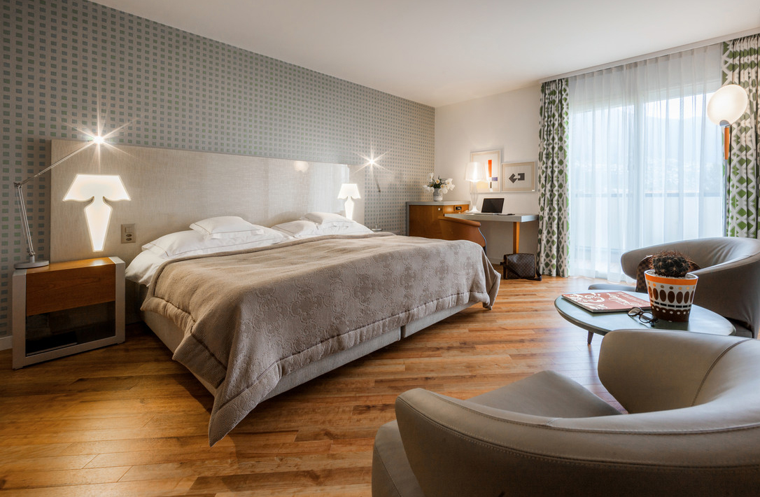 Modern hotel room with large double bed, cozy seating area, work desk, wooden flooring, graphic accent wall, and balcony with natural light.