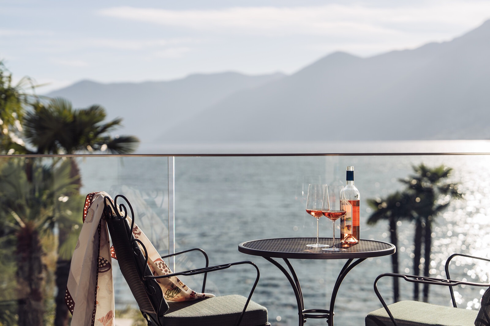 Balkonterrasse mit Roséwein, zwei Gläsern und Bergblick über einen See mit Palmen im Vordergrund.