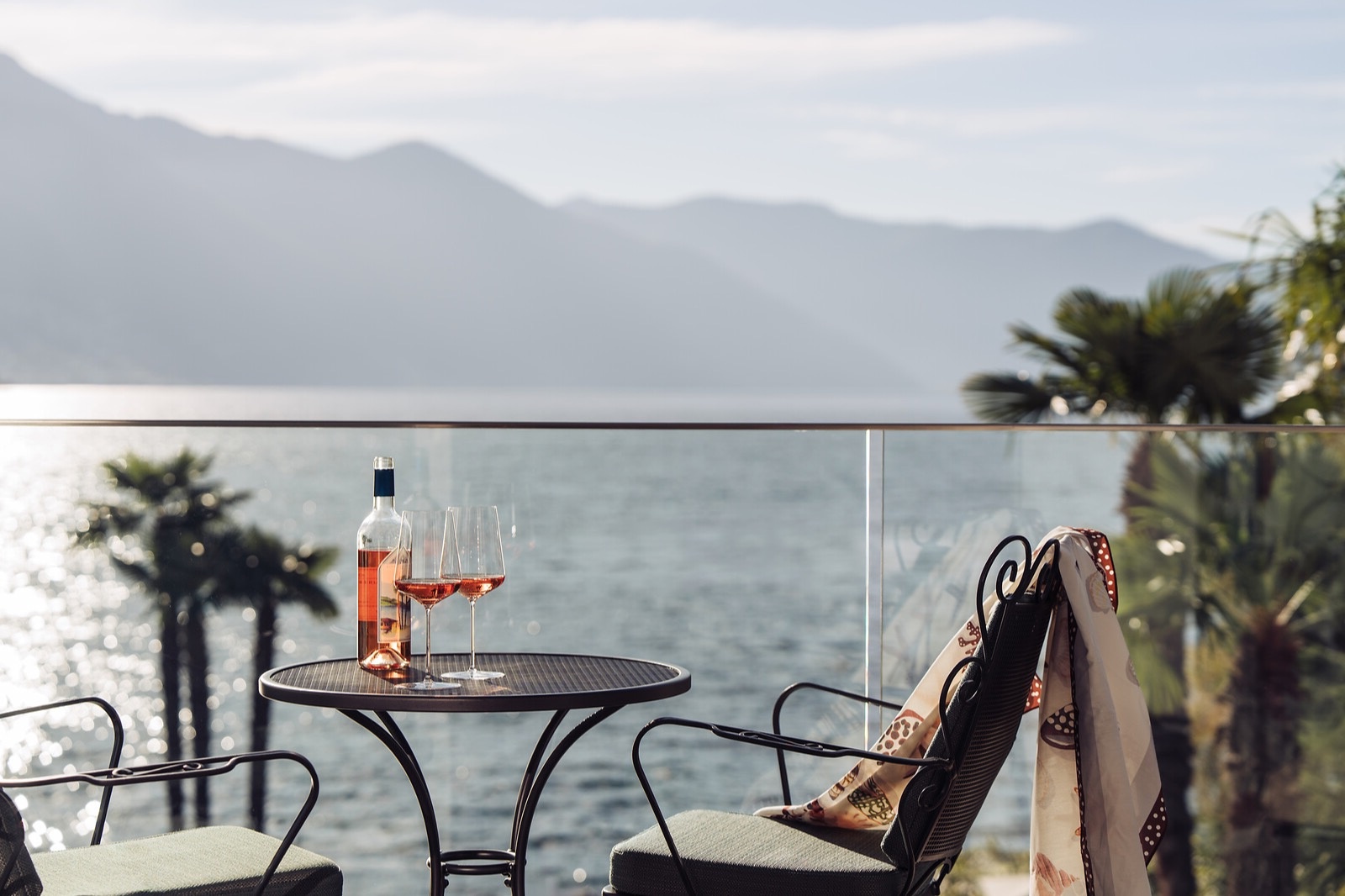 Balcony terrace with two chairs and table set with rose wine glasses overlooking a lake with mountains in background.