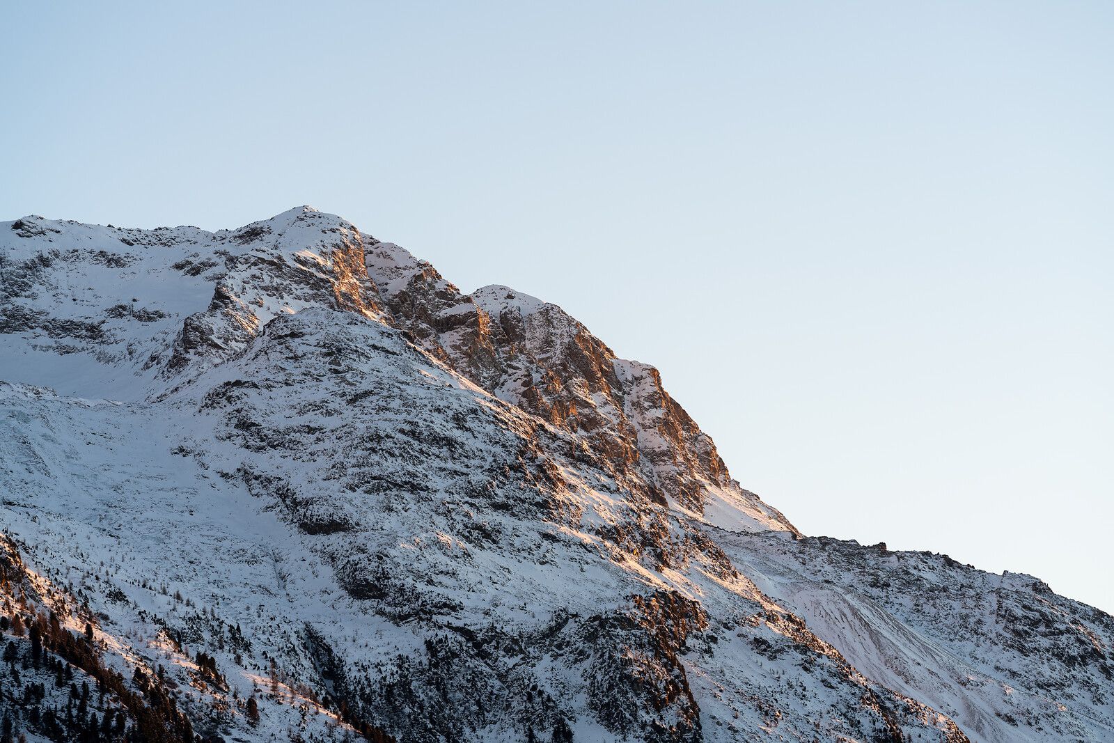 Schneebedeckter Berggipfel mit freiliegenden braunen Felswänden, die von goldenem Sonnenlicht beleuchtet werden, vor einem blassblauen Himmel.