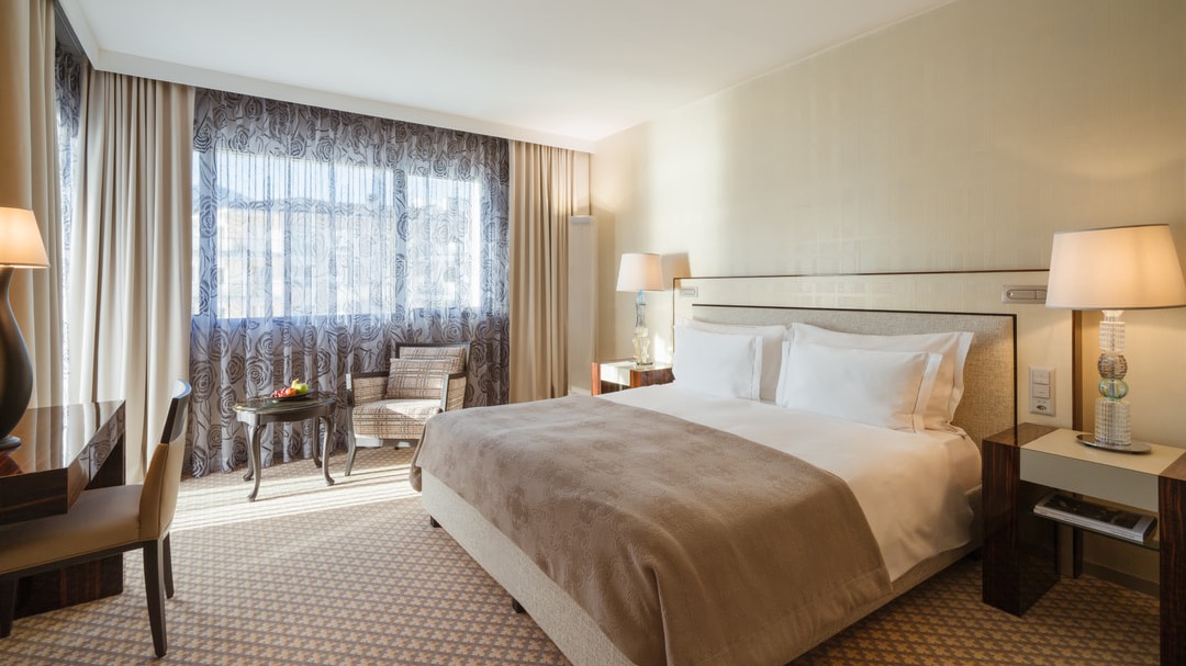 Großzügiges Doppelzimmer mit gemütlichem Sessel, Beistelltisch mit Obst, Tageslicht durch bodentiefe Fenster und dezentem Design.